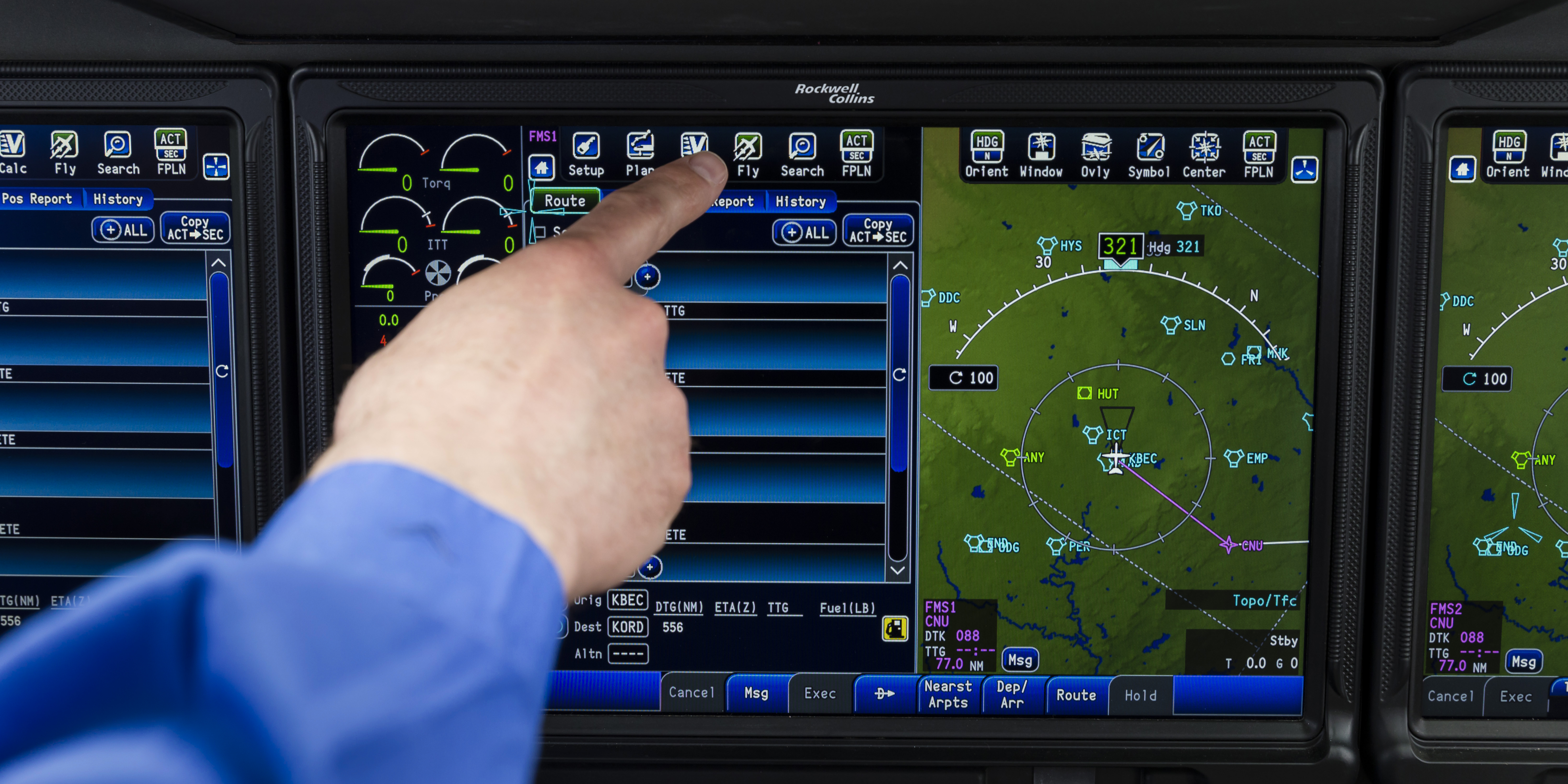 A close-up view of an aircraft cockpit touchscreen display showing navigation and flight management interfaces. A hand in a blue sleeve is pointing toward a button on the upper part of the center screen, which displays route options and system controls. The screen on the right shows a moving map with waypoints, terrain, and radar-like circular range indicators.